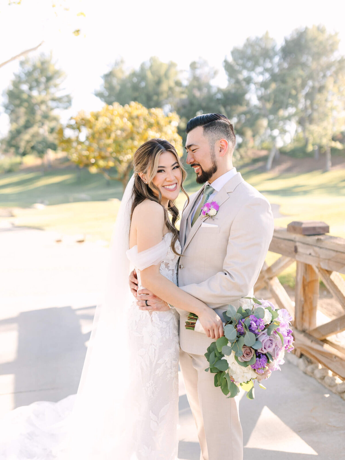 A joyful bride in a lace gown and groom in a beige suit embrace outdoors at Coyote Hills Golf Course. They're smiling, surrounded by green trees, with a vibrant bouquet.