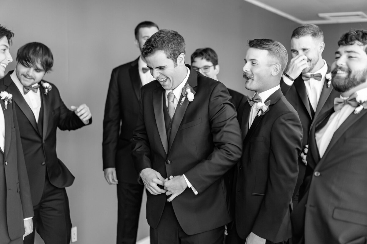 A group of men in suits and boutonnieres  stand together outdoors, laughing and smiling. The atmosphere appears joyful and celebratory. The photo is in black and white.