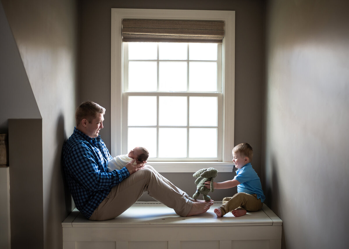 Dad sits in window with newborn as older brother plays with his stuffed rabbit.