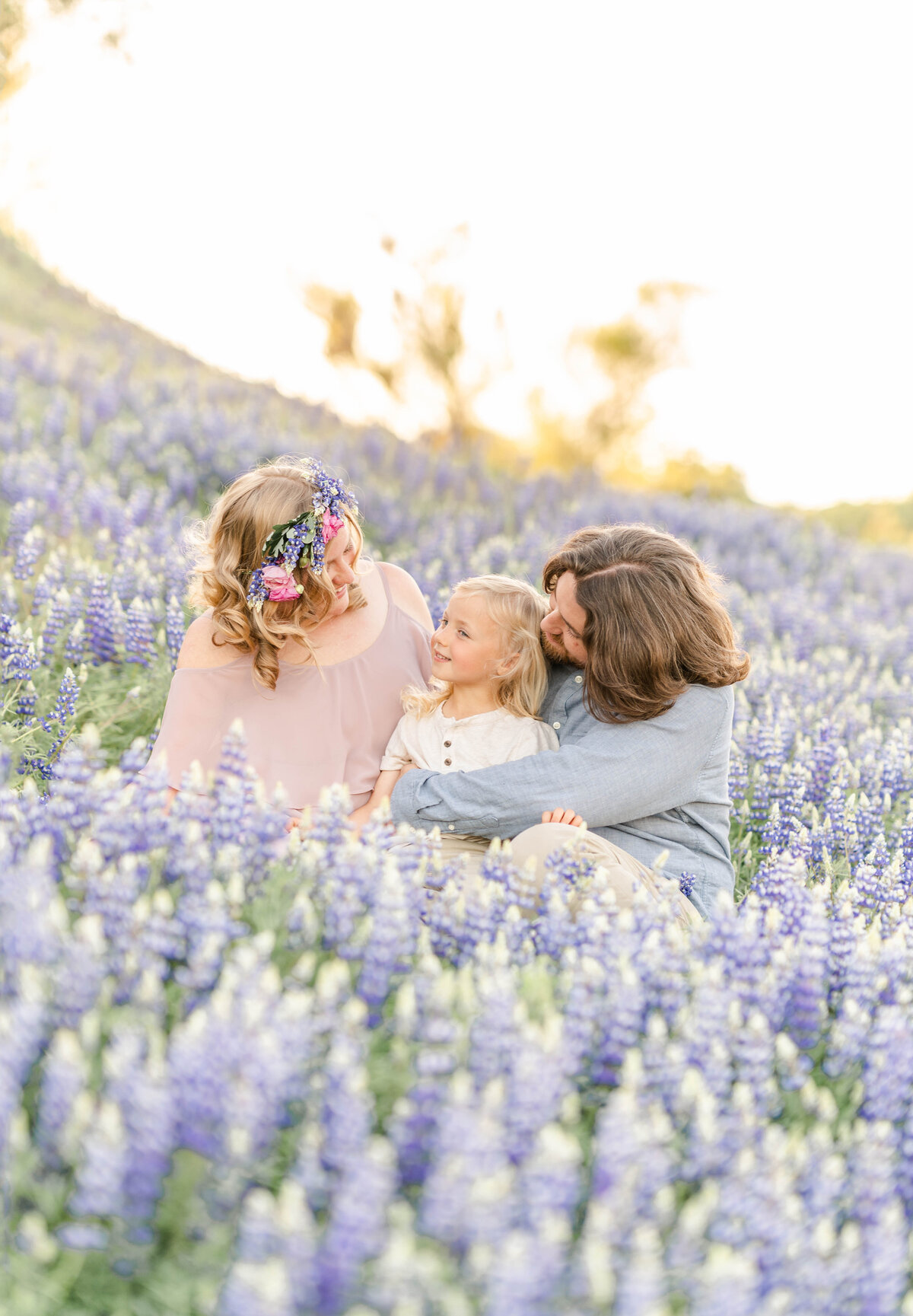 A family of three sits in a field of purple lupines hugging and smiling while the mother wears a floral headpiece, photographed by Bay Area Photographer, Light Livin Photography.