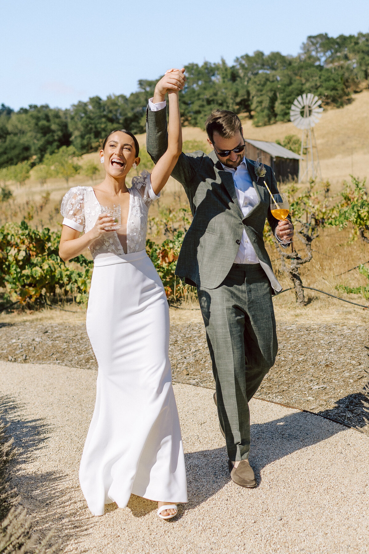 Celebrate love and happiness with a couple's joyful stroll through a scenic vineyard. Featuring the bride in a stylish gown with lace details and the groom in a dapper suit, this image is perfect inspiration for couples seeking a vibrant and elegant outdoor wedding.