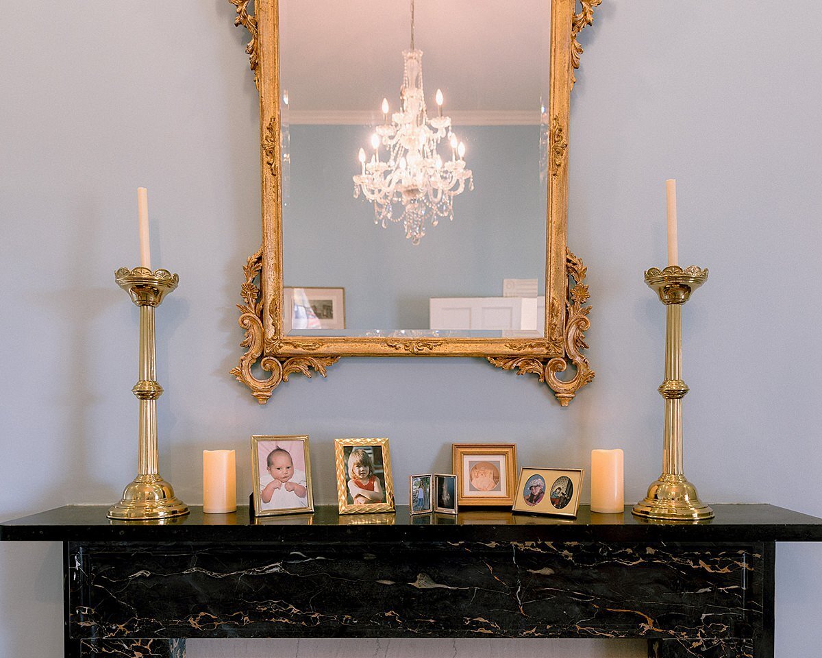 A black cast iron mantle against a light blue wall with a gold framed mirror and tall gold candle sticks with white taper candles stand on either side of framed family photos. A crystal chandelier reflects in the ornate gold framed mirror at Ravenswood Mansion