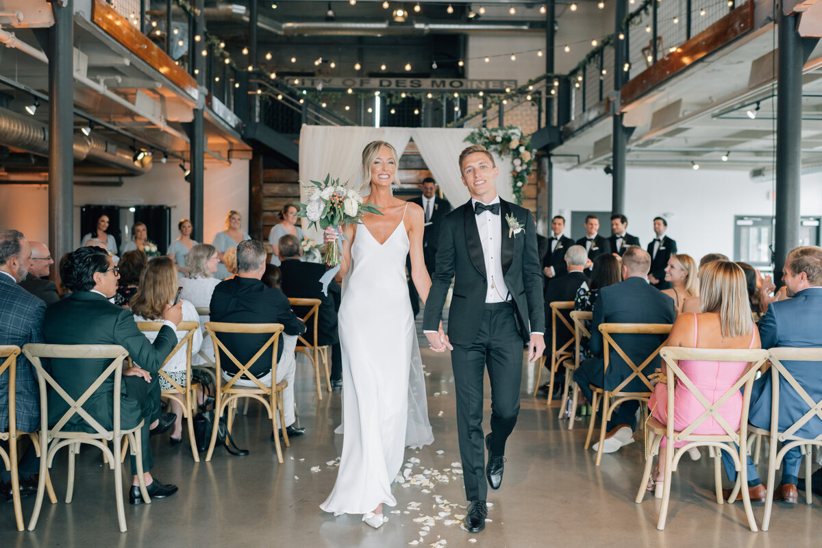 Bride wearing a chic wedding gown and groom wearing a black tuxedo share a kiss in a tunnel of people at The River Center. Photo by Omaha Wedding Photographer, Anna Brace