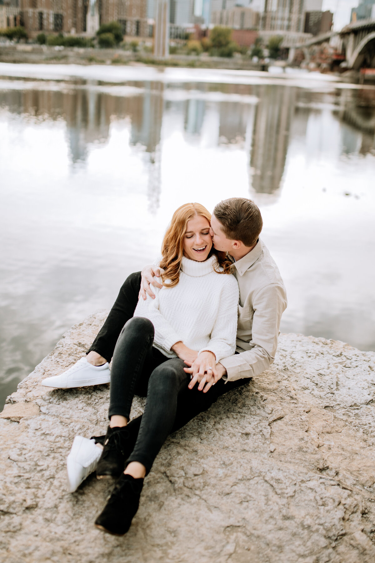 Casual couple sits upon a rock along the Mississippi River, as the iconic Stone Arch Bridge towers in the background during a Minneapolis engagement photoshoot.