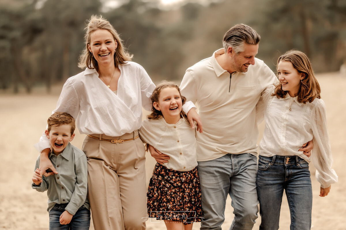Gezinsfoto van mama, papa, twee dochters en 1 zoon die allemaal elkaar vasthouden op een lijn en lachend en kijkent naast elkaar lopen in de duinen van Herperduin