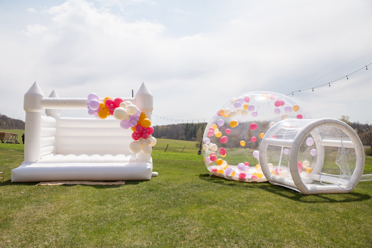 A white bounce house with a colorful balloon garland on the front, and a giant bubble dome next to it with balloons inside.