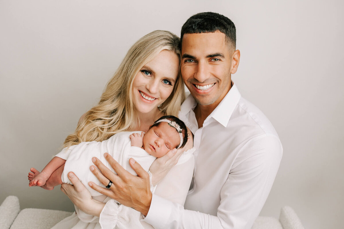 mom with blonde hair and dad with brown hair smiling at the camera and holding newborn daugher who is wrapped in white