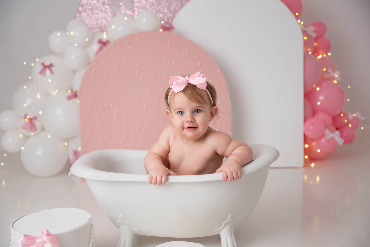 Baby leaning over with both hands on the edge of a mini tub, surrounded by pink and white balloon arches and pink bows.