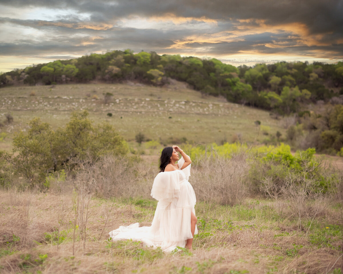 Pregnant woman in flowing white gown sitting on hillside during golden hour maternity photography session in Austin