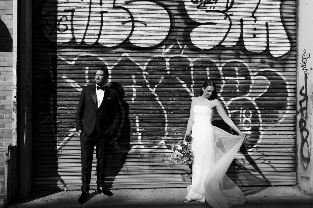 black and white photo of a bride and groom standing in front of a graffiti wall while she holds up the side of her dress