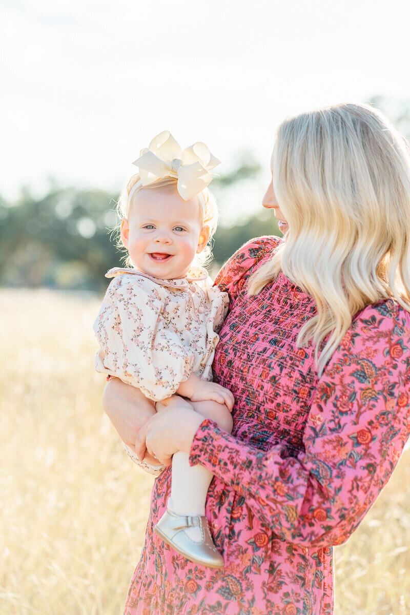 a mother holds her toddler girl in her arms and looks at her while her daughter looks at the camera of their Austin family photographer.