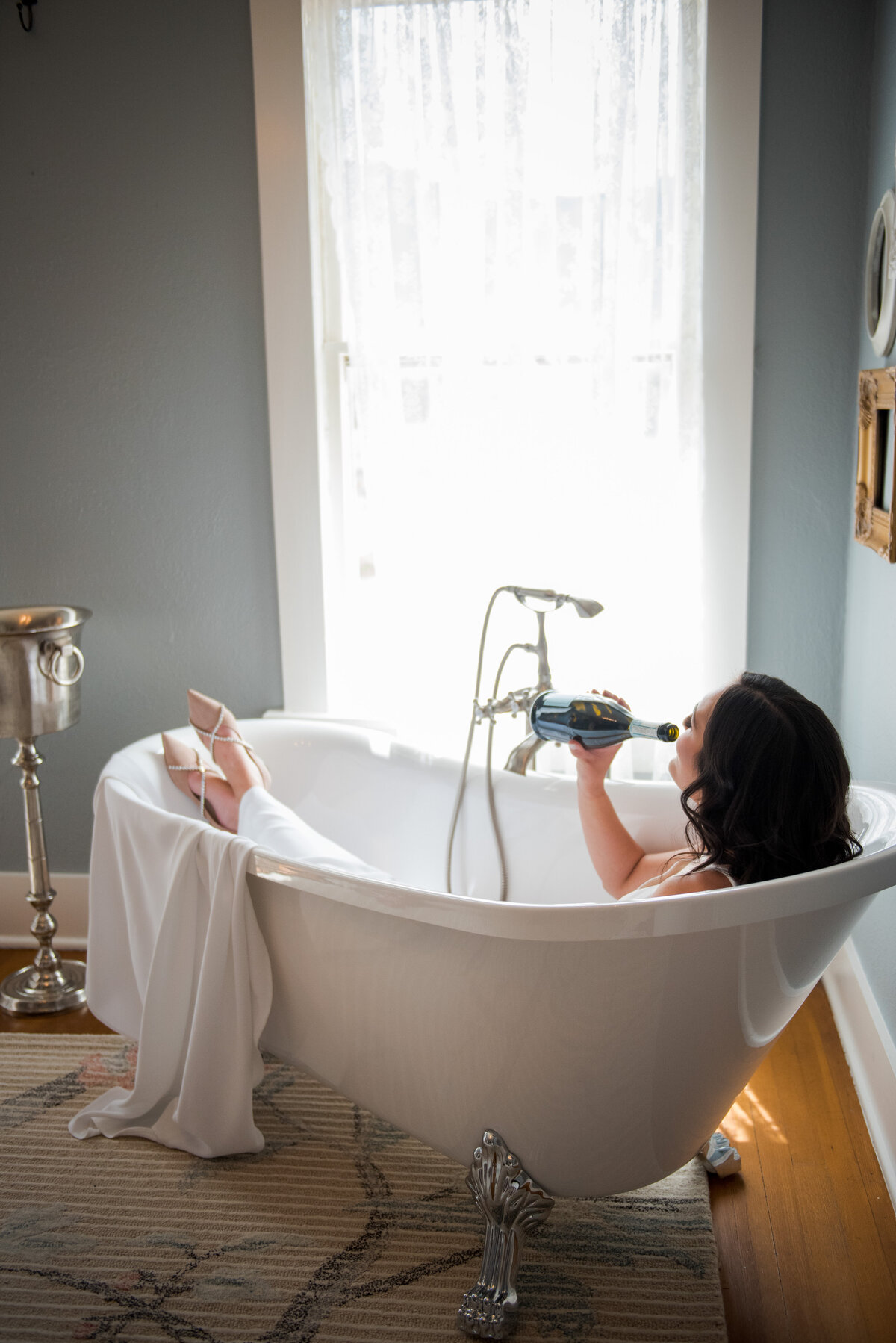 A playful shot of a bride sitting in a clawfoot bathtub in her wedding dress while sipping a bottle of champagne.