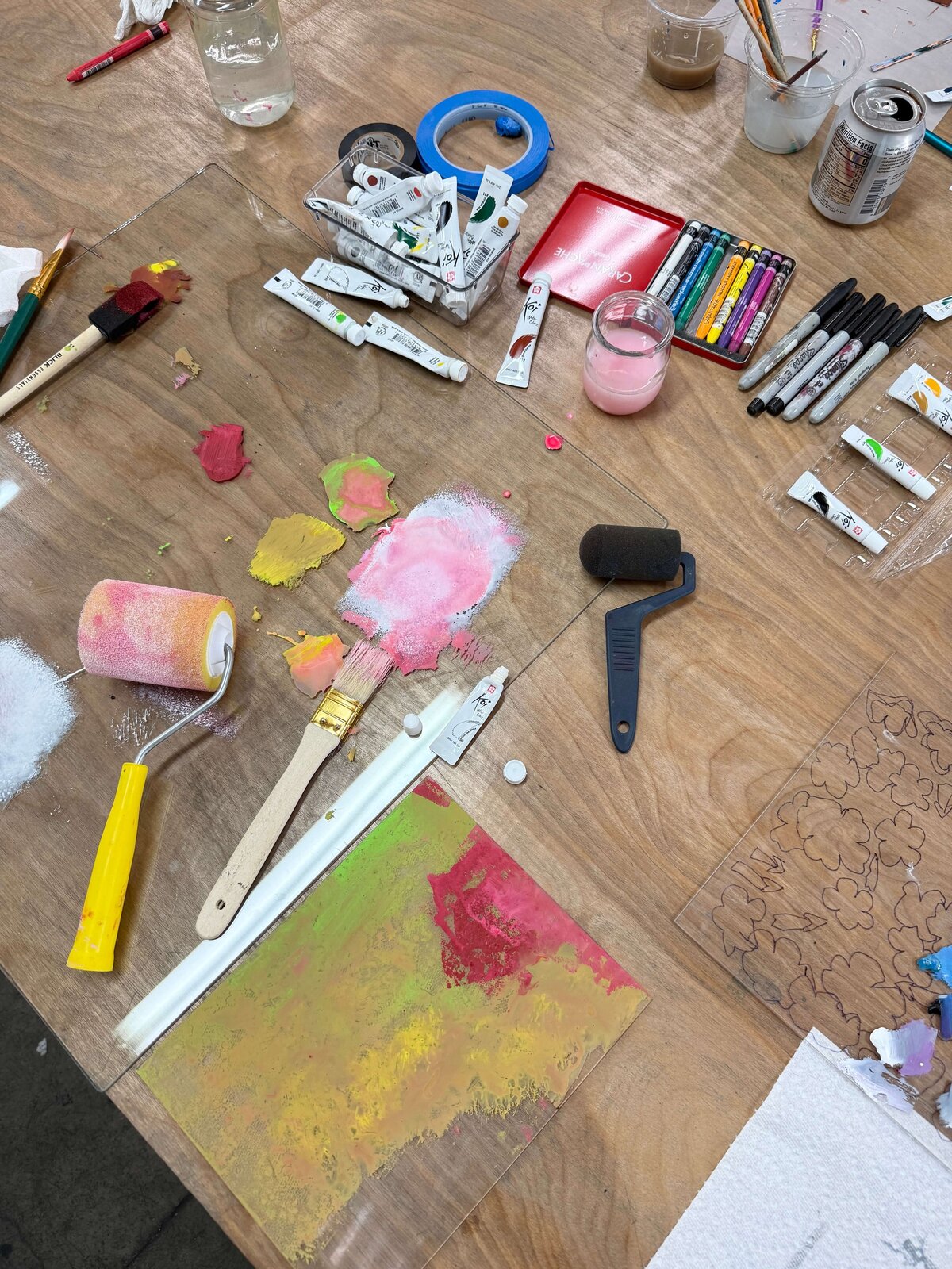 A closeup of the studio table showing a roller, brushes, pigments, and a vibrant inked plate during the monoprinting process.