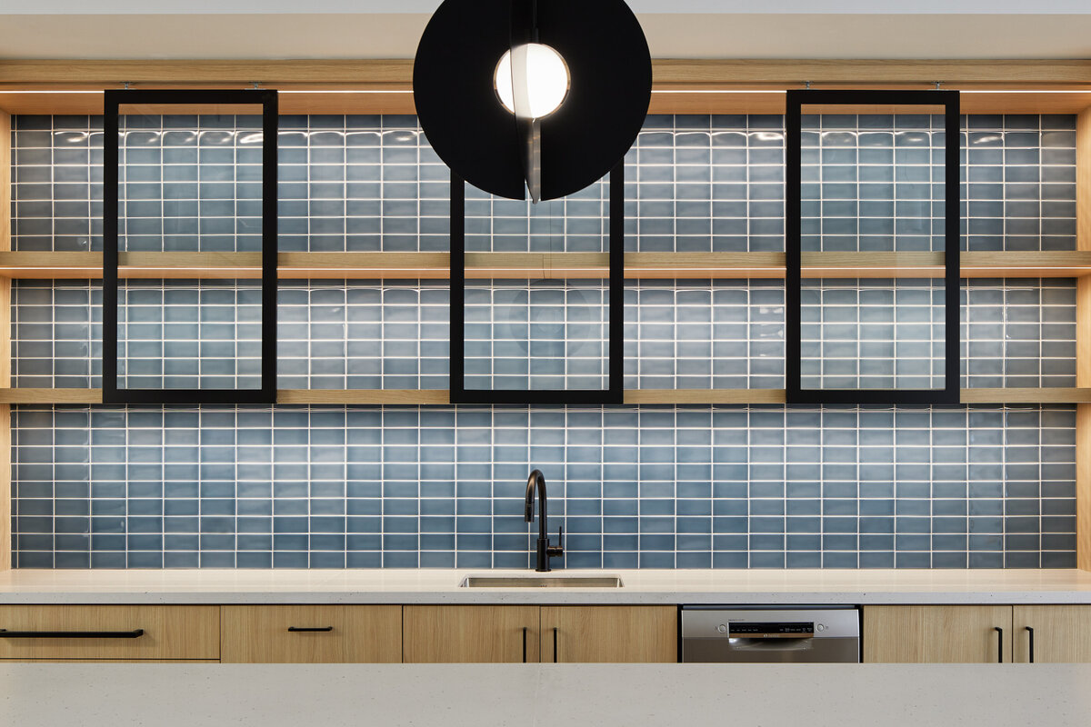 A kitchen counter and sink area featuring a blue tile backsplash, wood cabinetry with black accents, open shelving with movable glass panels, and an Orbit pendant light