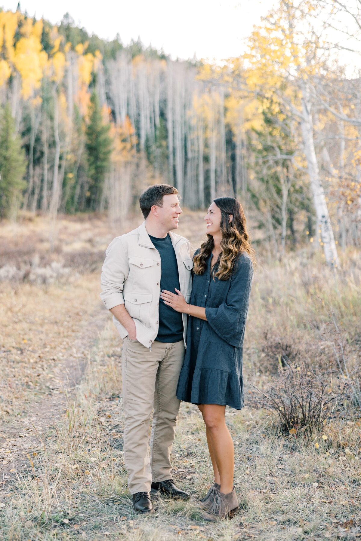 This is a portrait of the young couple. They are looking at one another surrounded by yellow aspen trees and mountain peaks. 