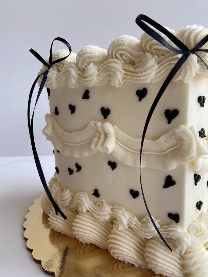 A Close up shot of the side of a vintage heart cake. Shot focuses on the layers of white piping, the tiny black hearts scattered all over, and the black ribbon accents.