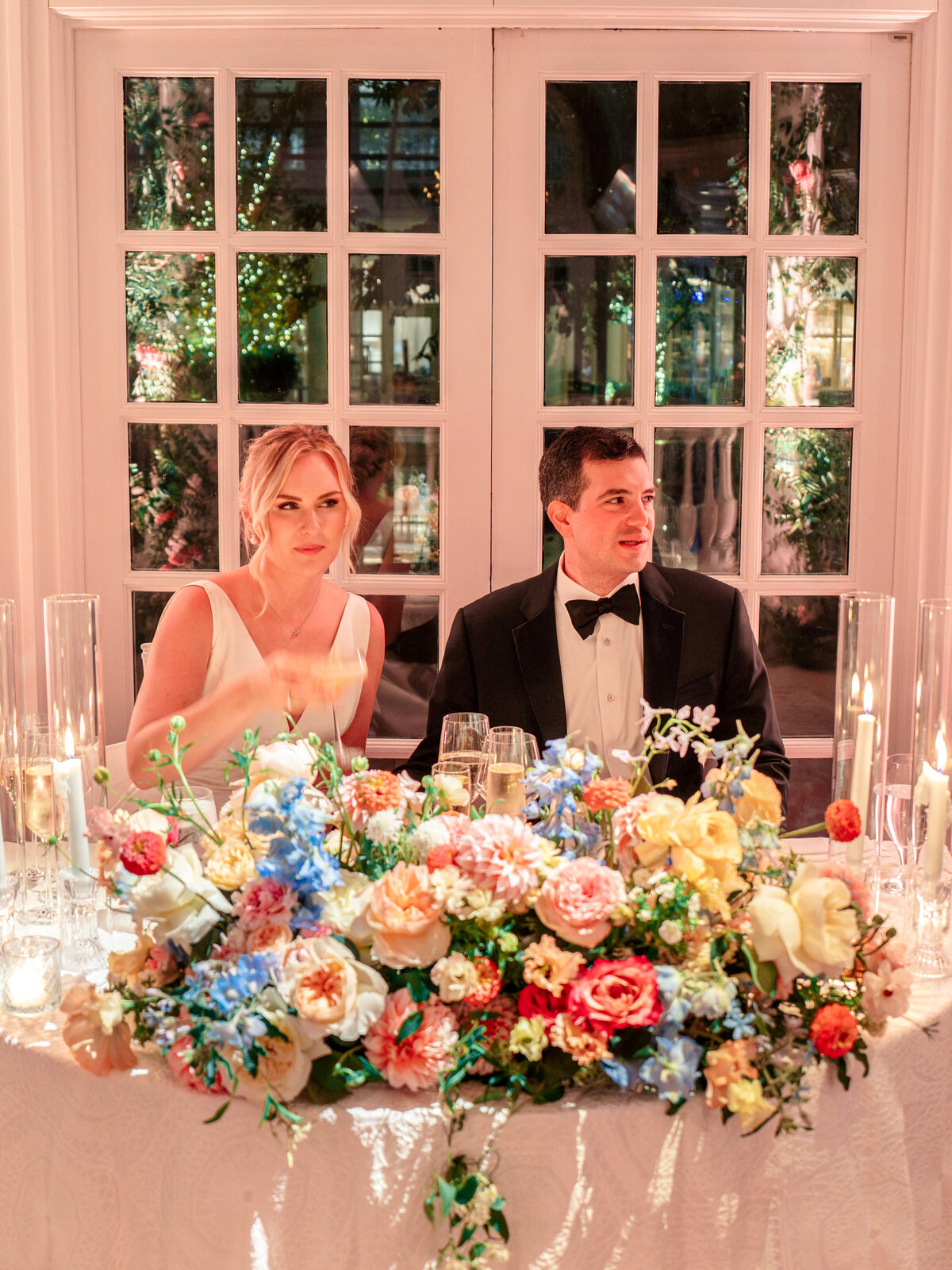 A couple sits at an elegantly decorated wedding table. The woman is in a white gown, and the man is in a black tuxedo. The table is adorned with colorful flowers and lit candles, with a large window behind them showing a garden with twinkling lights.