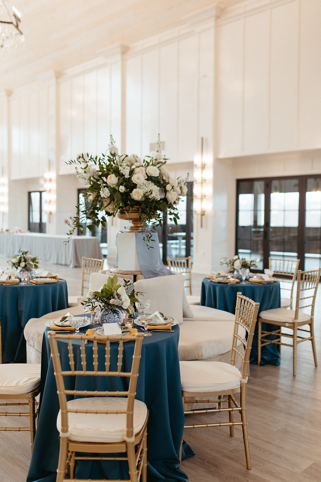 A bright and airy banquet room with blue satin linens on round tables, gold chairs and white floral arrangements.