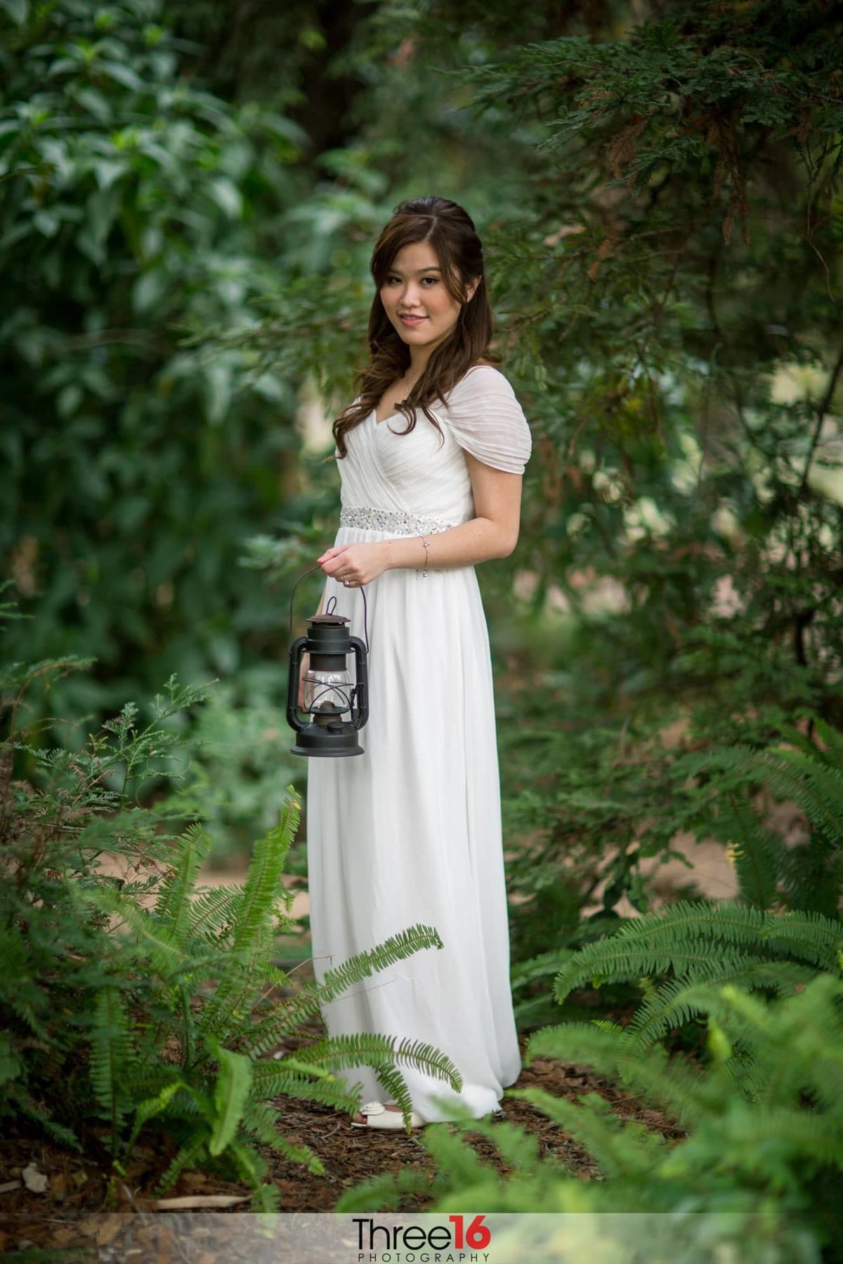 Bride poses for photos amongst the green shrubs of the Los Angeles Arboretum holding a train-style lamp