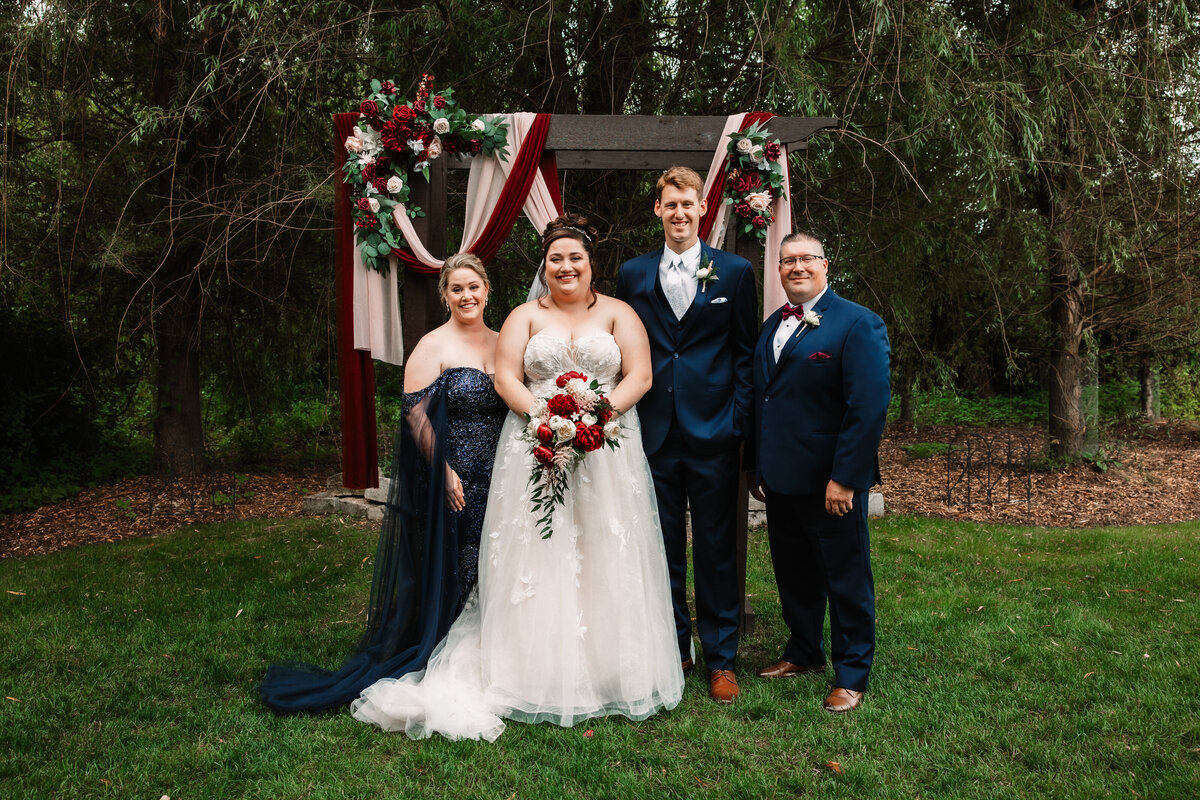 Newlyweds stand with best man and maid of honor under the arbor smiling