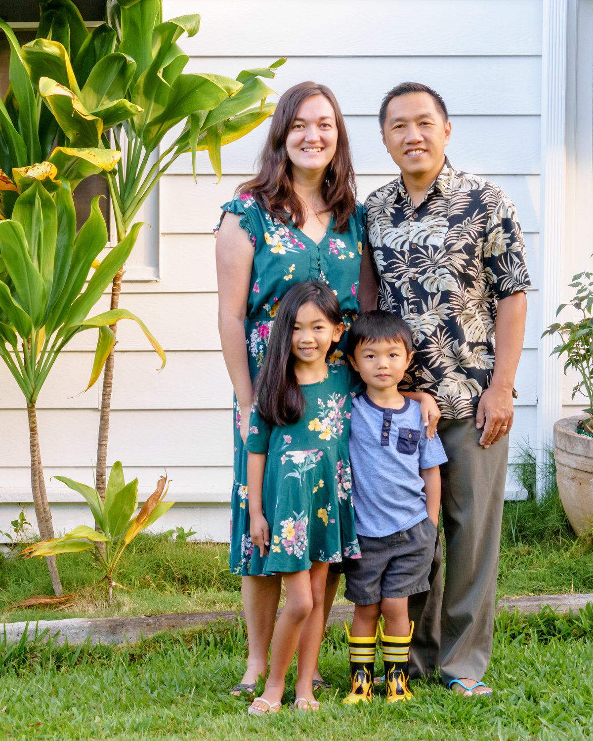 Wife and daughter wearing green floral dresses along with husband and son stand in front of a white house