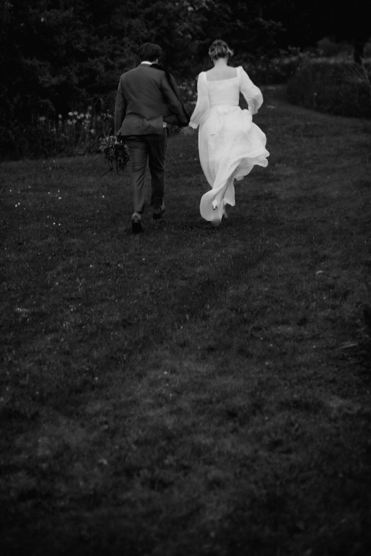 Das Hochzeitspaar rennt schwungvoll über eine Wiese. Die Kamera ist von hinten auf sie gerichtet.