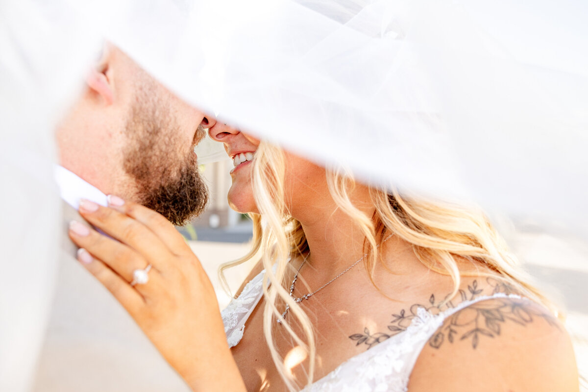 The bride and groom share a romantic moment under the bride’s flowing veil, their faces inches apart as they smile at each other.. Captured by Cassie Lee Photography