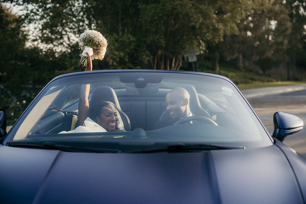 Bride and groom sit in the wedding car smiling. Bride holds up her wedding bouquet.