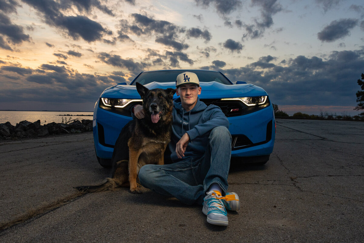 A senior guy sitting on the ground with his dog in front of his car during golden hour in Lawrence, KS