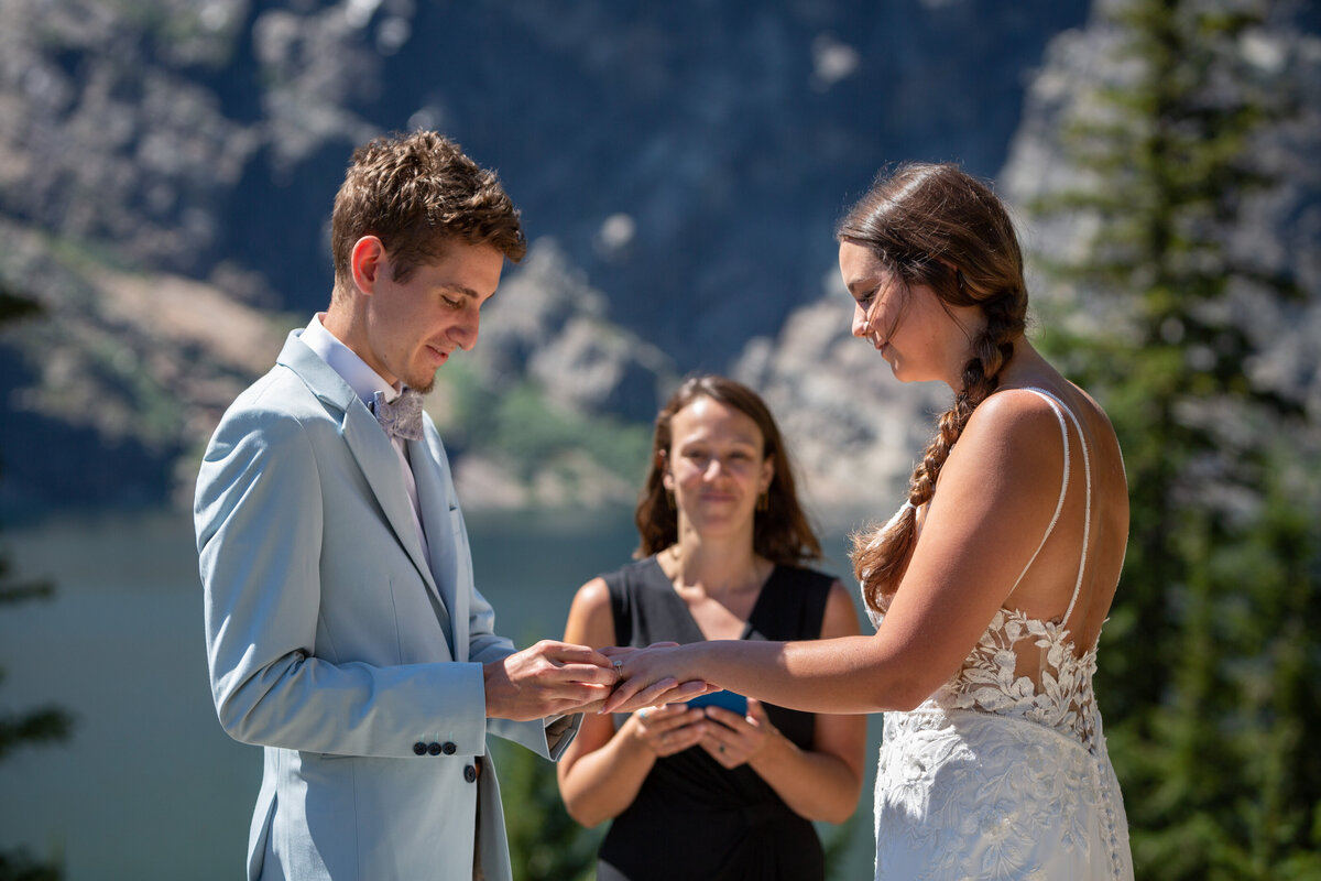 A groom slides the wedding ring on his bride's finger during their elopement ceremony and their officiant smiles behind them.