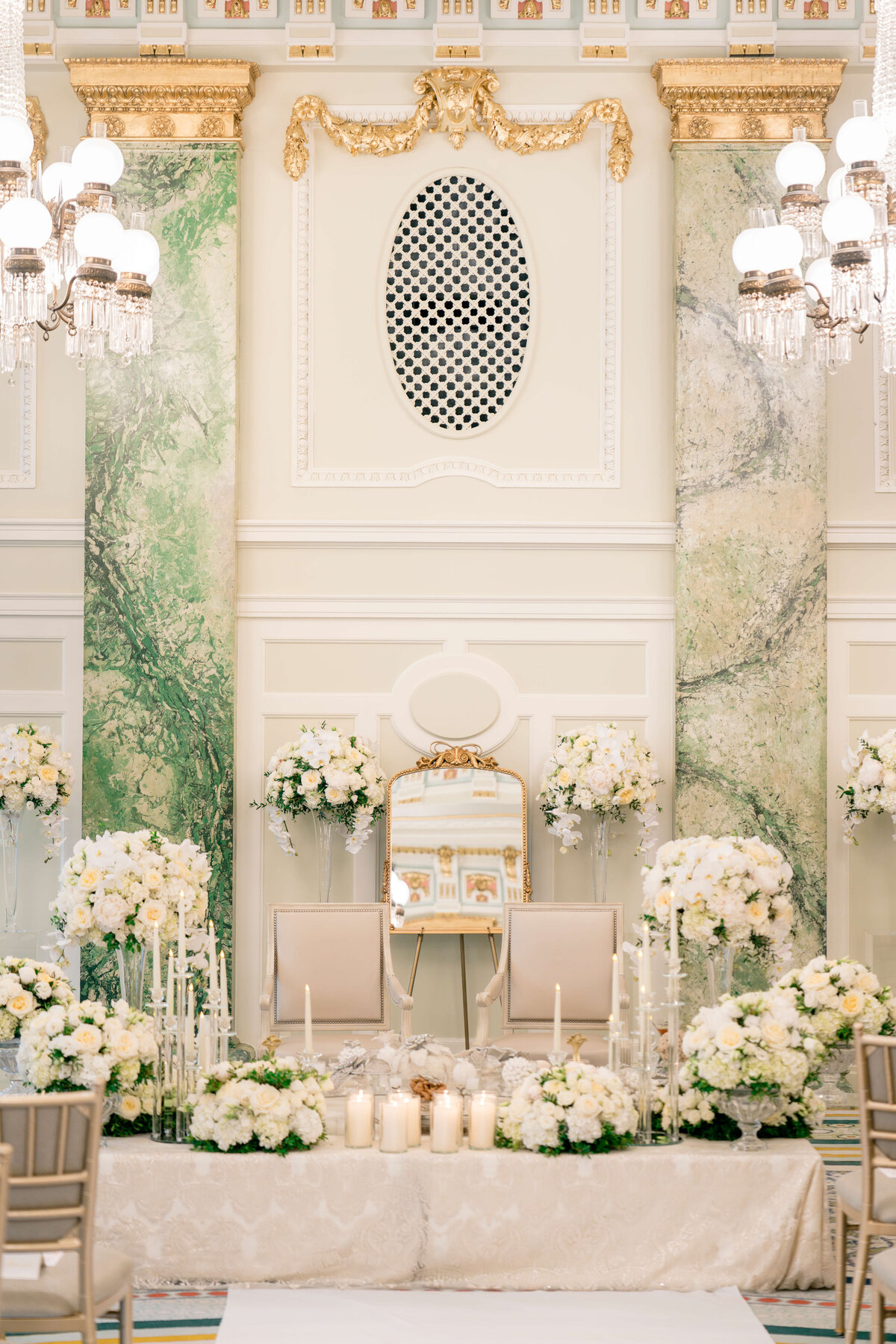 A lavish wedding altar is adorned with white and cream flowers, candles, and elegant chandeliers. The backdrop features ornate gold detailing and a green marble design. Two tall chairs are positioned at the center.