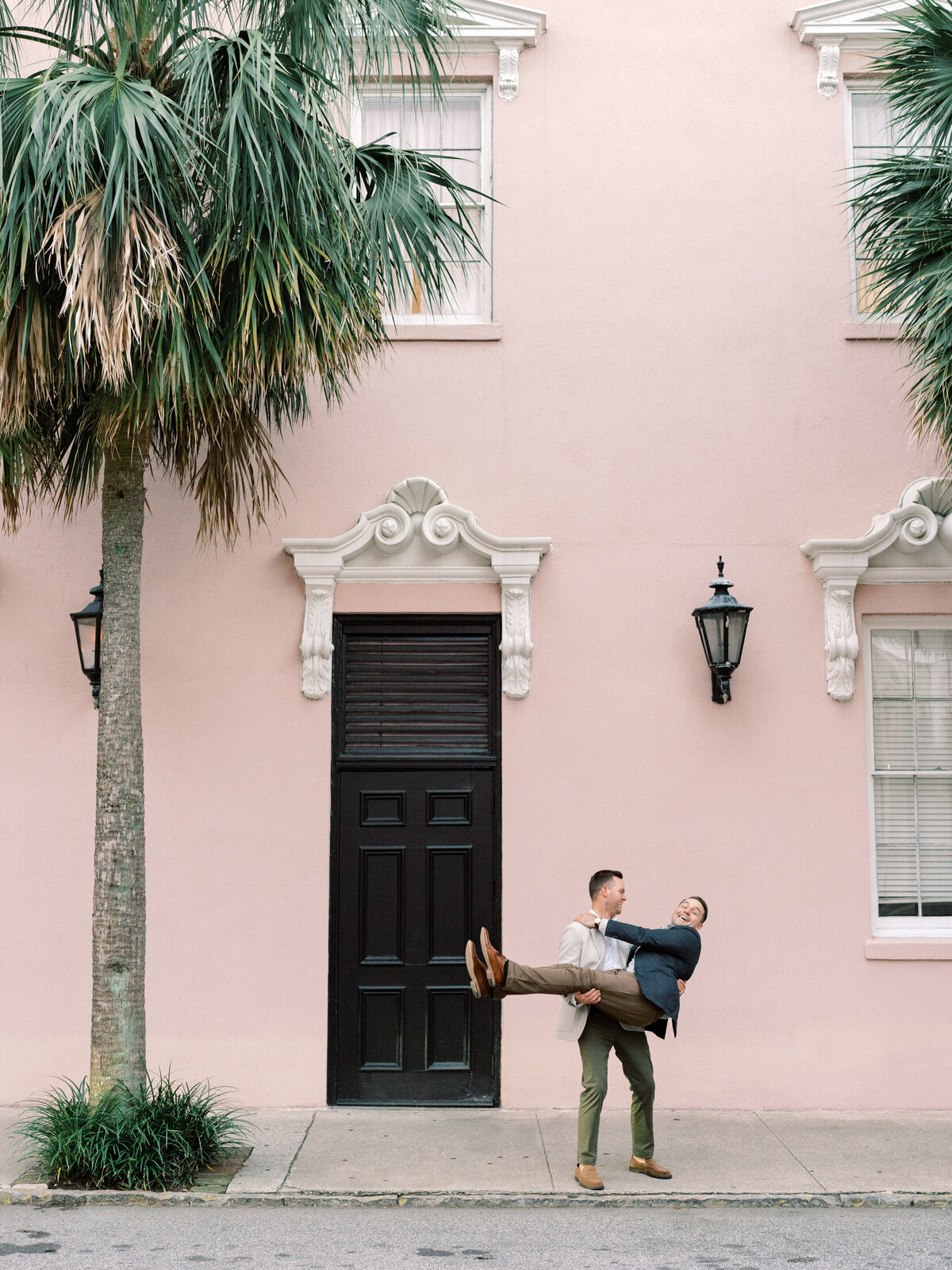 In a playful moment, one partner lifts the other, showcasing a blend of affection and fun, captured against the pastel Mill's House Hotel in Charleston, accentuating their joyful companionship.