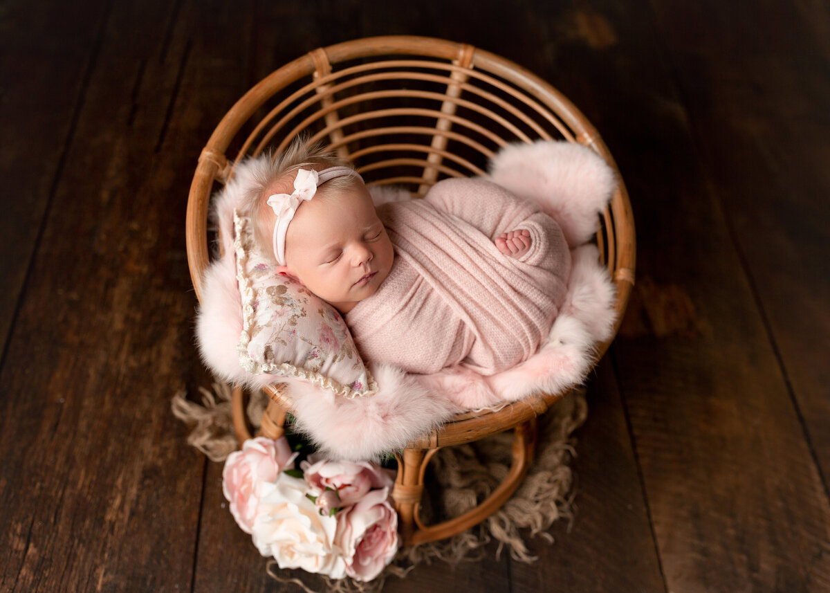 Baby girl sleeping on a miniature rattan chair for newborn photoshoot. Baby is wrapped in a pink knit with her toes peeking out. She is resting on a pink fuzzy rug and resting her head on a pink and cream floral pillow.