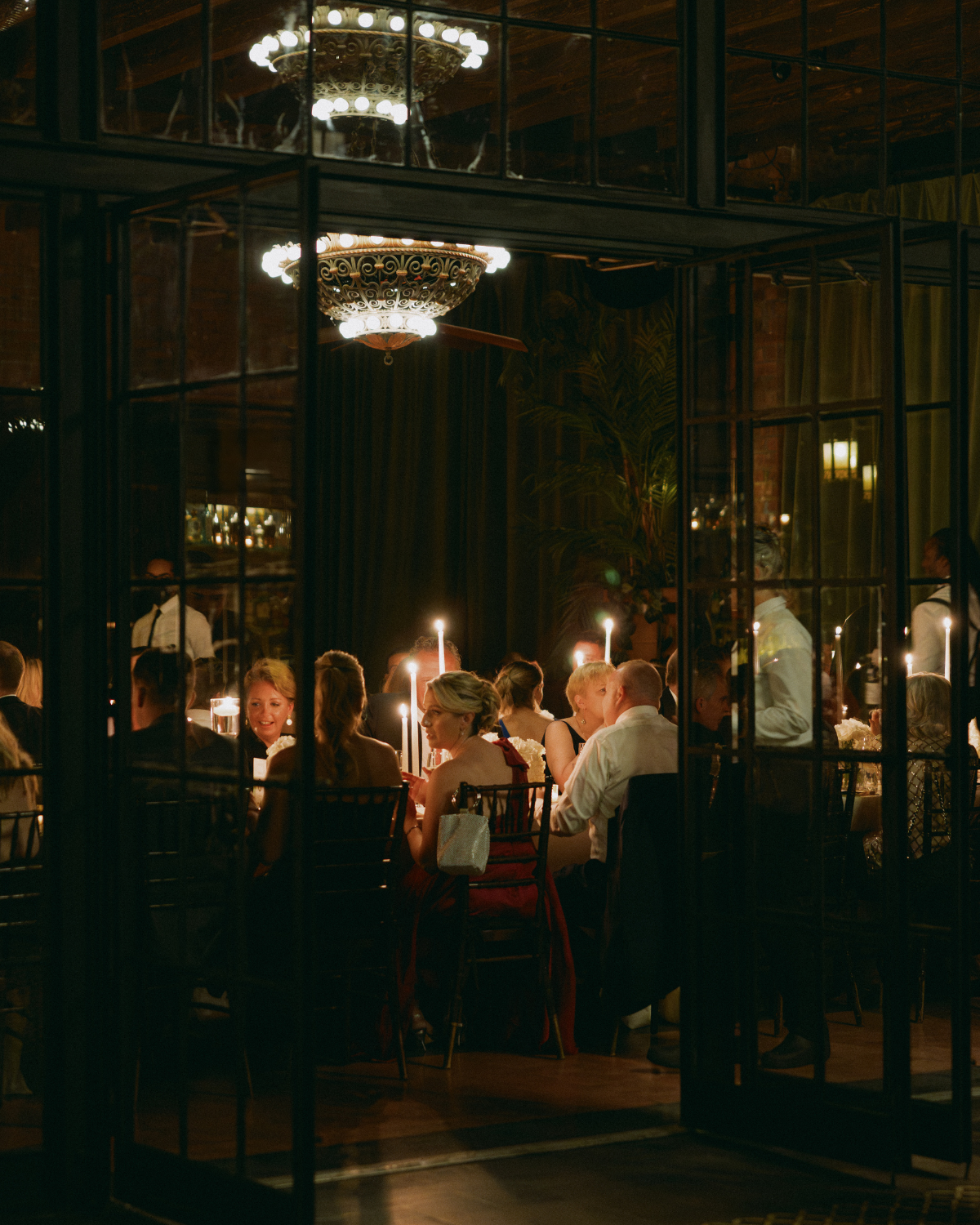 Guests seated at an elegant candlelit wedding reception in New York City, viewed through glass doors with a vintage chandelier glowing above, capturing a romantic and intimate evening celebration