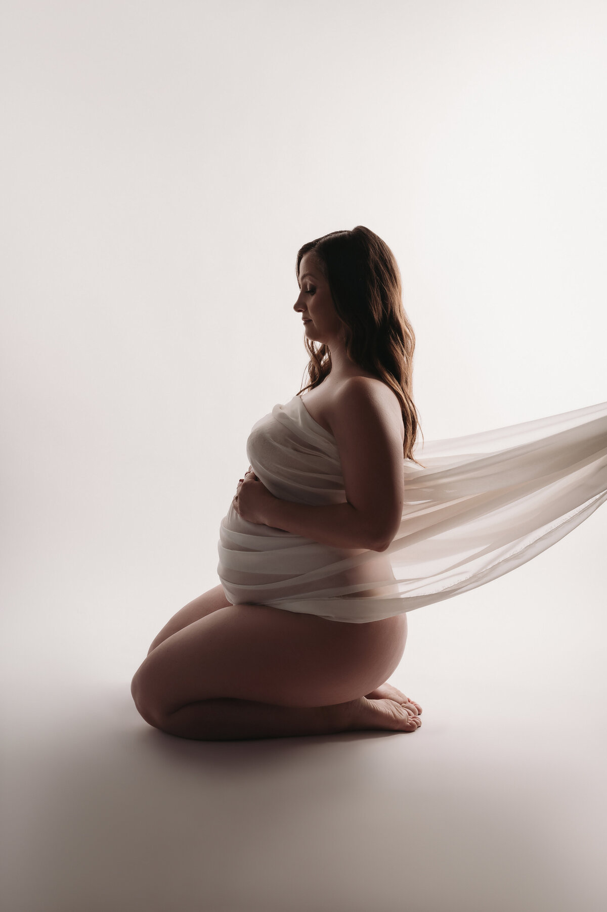silhouette maternity  of woman kneeling  side profile with  white pearl fabric hugging her pregnant bump