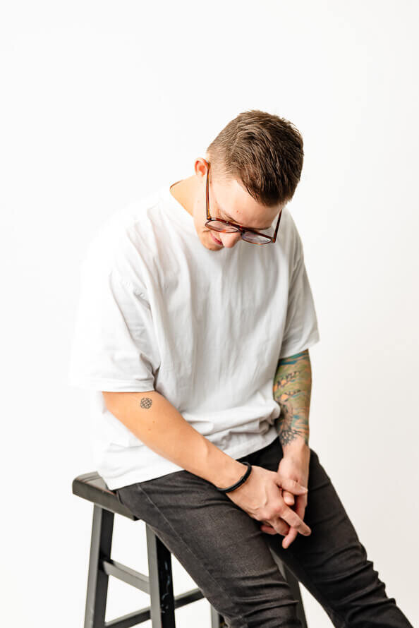 Man sits on stool in front of white background and looks down.