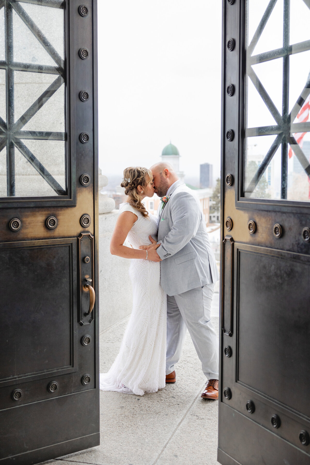 A couple shares a kiss framed by the elegant black iron doors of the Utah State Capitol, with the cityscape visible in the background