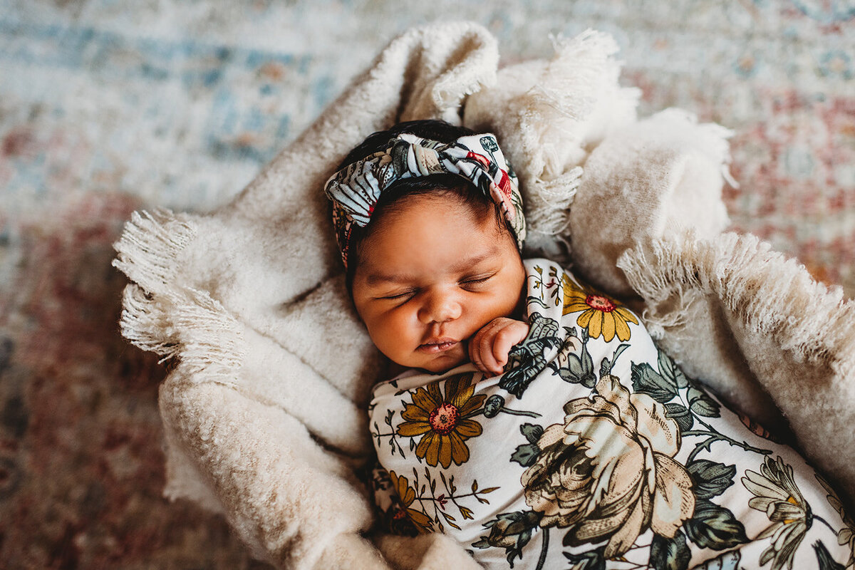 newborn baby girl pictures with baby in a floral swaddle and bow laying in a basket with a fluffy white blanket on a run captured by Family photographers Maryland