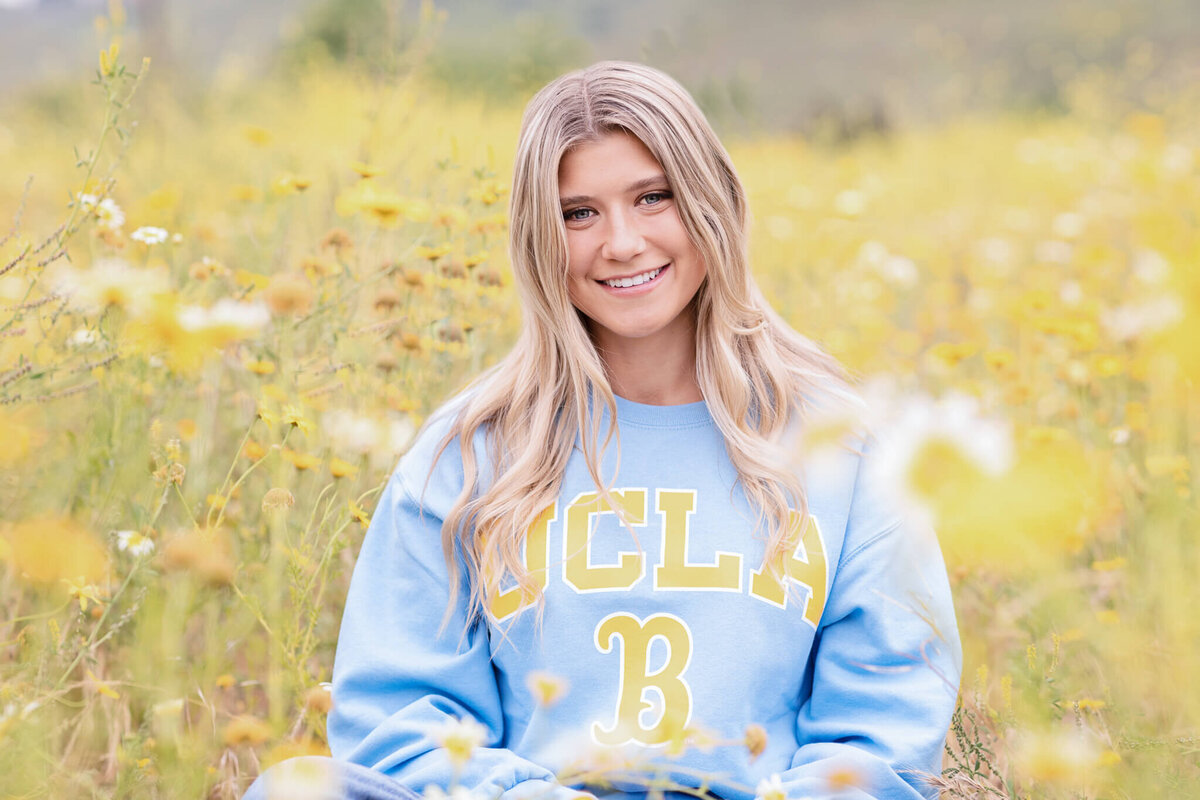 A person with long hair sits in a field of yellow wildflowers, smiling. They wear a light blue UCLA sweatshirt. The background is blurred, giving a soft, natural feel to the image.