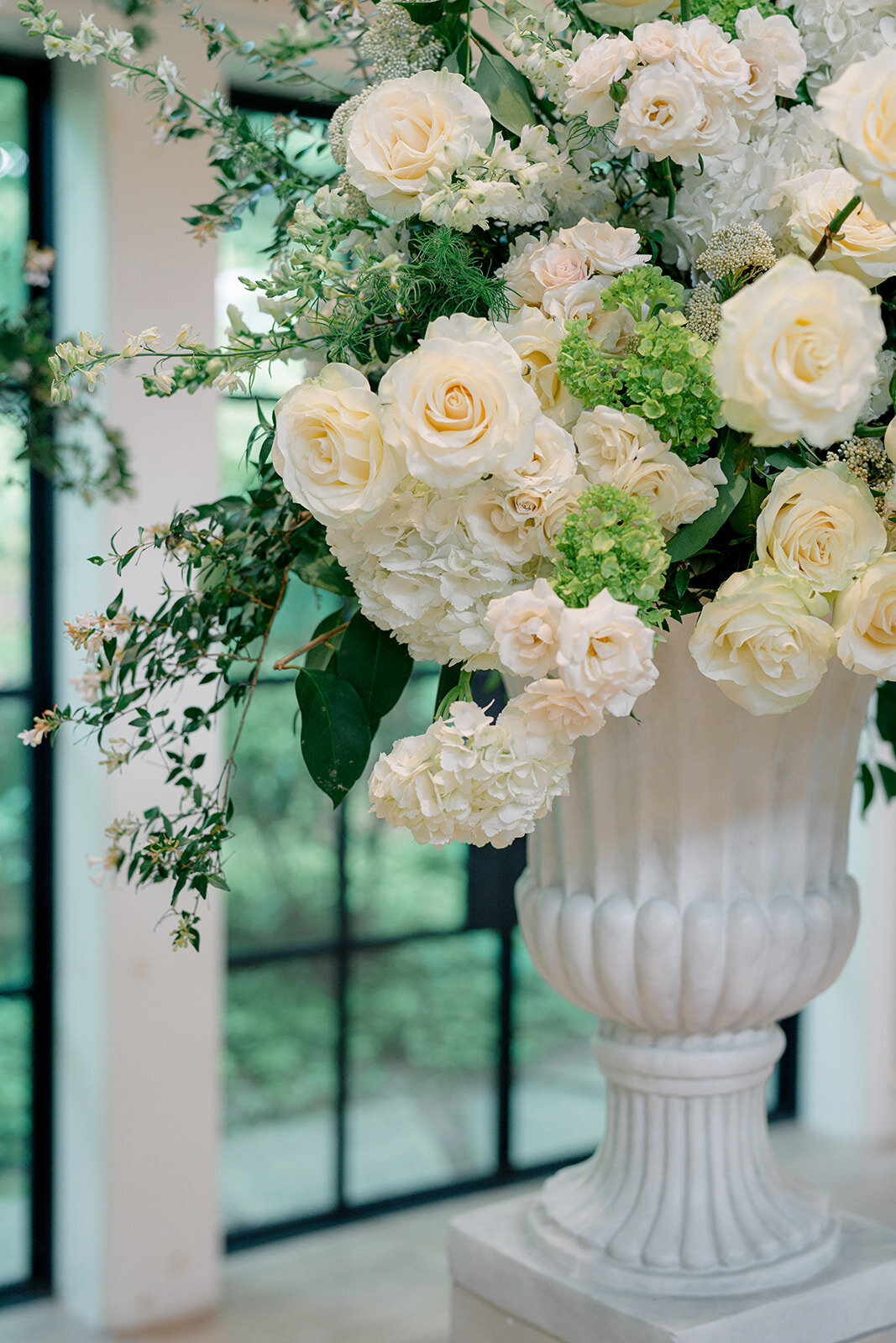 Statement floral urns filled with white roses and greenery at indoor wedding ceremony.”