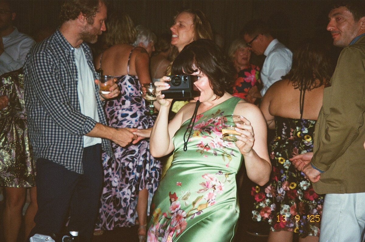 Woman in bright colored wedding guest dress snaps a photo of a bride and groom in upstate New York. 