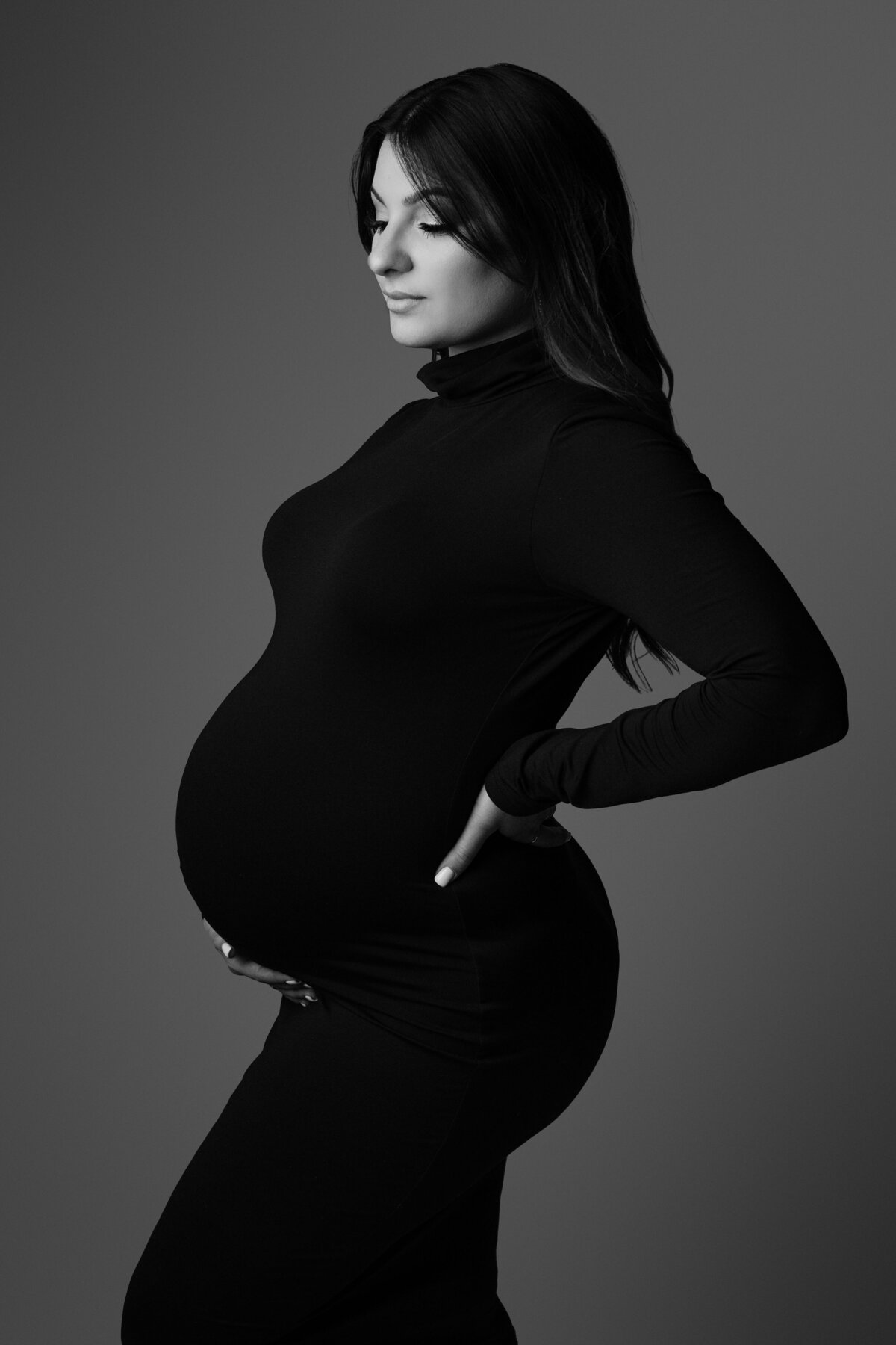 Black and white maternity portrait of a woman wearing long sleeve fitted black long dress holding one hand behind back and other hand on tummy looking down at baby bump
