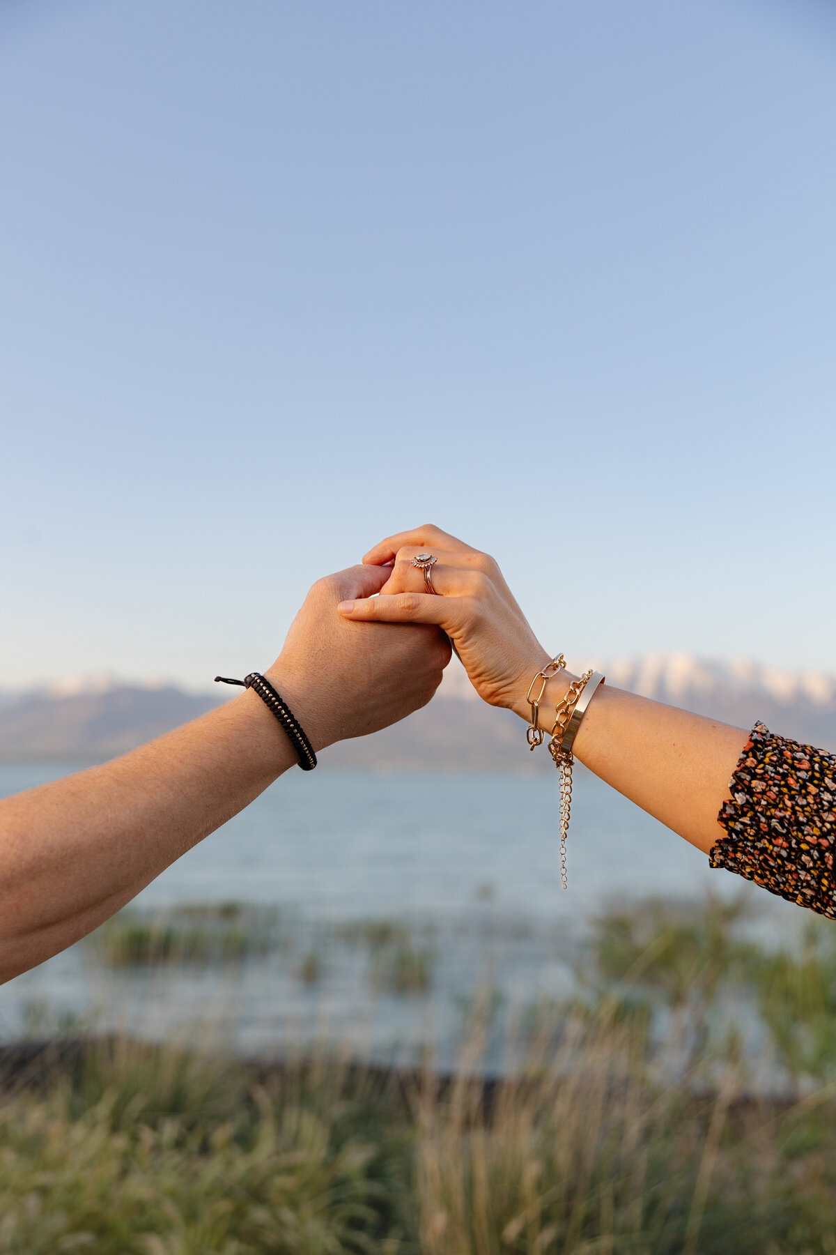 A close-up of a couple’s hands clasped together, showcasing the woman's engagement ring against the backdrop of Utah Lake and distant mountains.