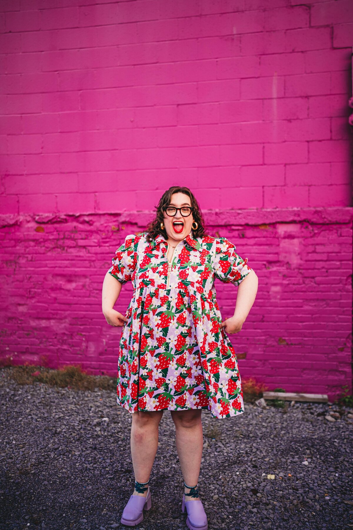 A joyful person in a strawberry dress poses in front of a bright pink wall, embodying confidence & color in an Erie, PA branding session.