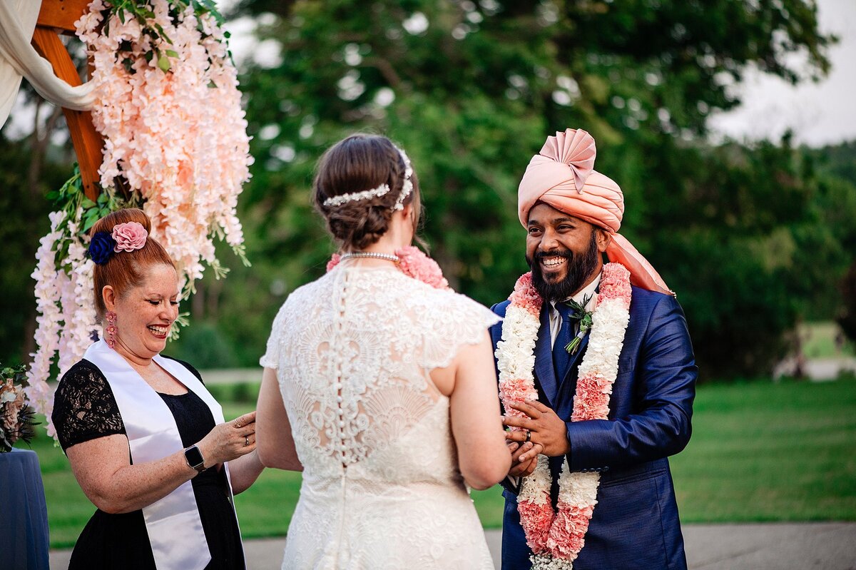 The groom, wearing a cobalt blue suit, pink turban and white and pink varmala garland smiles at the bride as they exchange vows in front of their officiant. the bride is wearing a lace wedding dress with capped sleeves and a rhinestone clip in her updo. The officiant stands to the left smiling. She is wearing a black lace dress with a white satin shawl and a pink flower in her hair. Behind her is a cedar wood arbor with hanging white drapery and large clusters of hanging pink flowers.