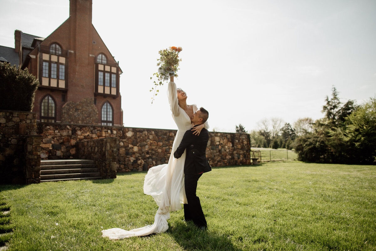 Wedding Photographer, a groom lifts his bride in the air, she lifts her wedding bouquet high on an estate grounds