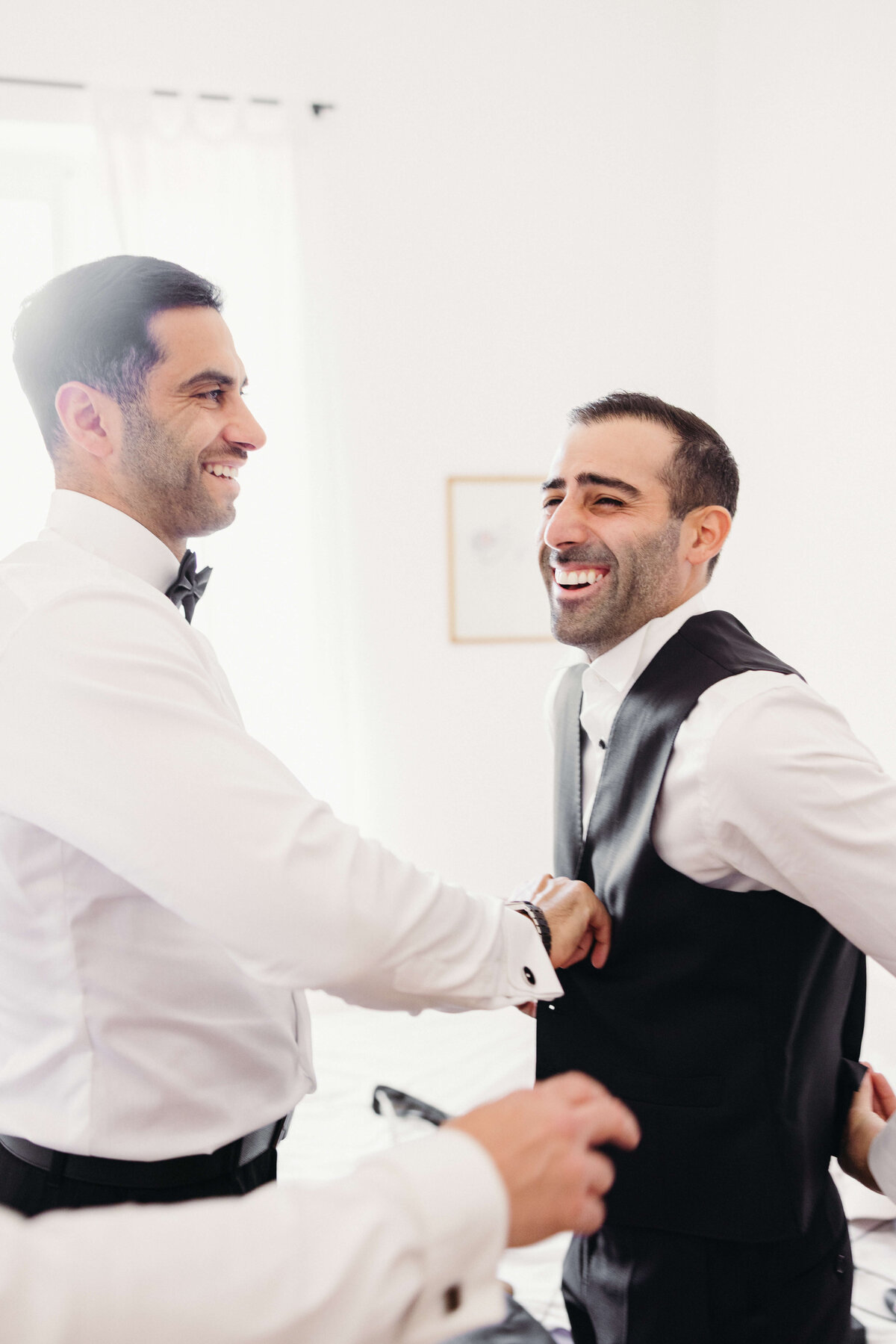 Groomsmen laughing while helping groom into vest before wedding

