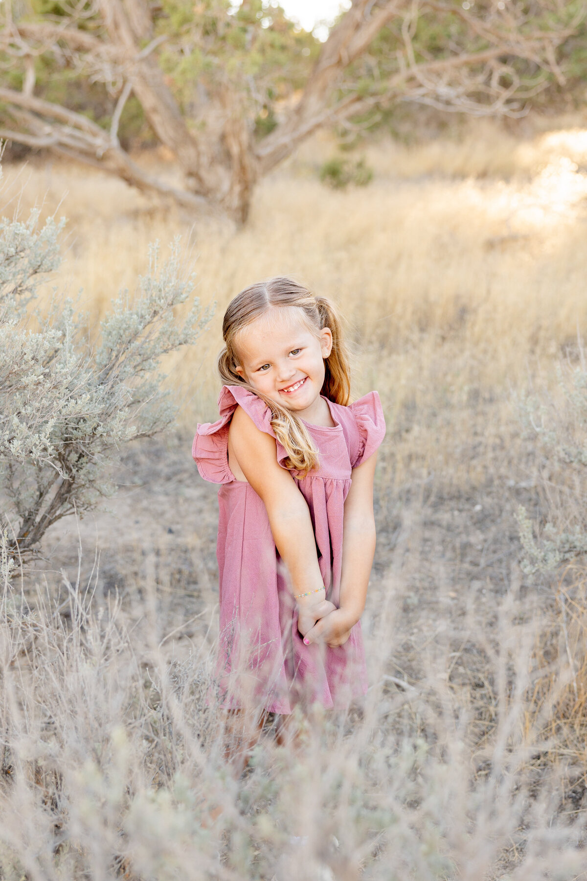 A young girl in a pink dress and pigtails stands among the tall grasses, her hands clasped in front of her as she gives a bright, playful smile.