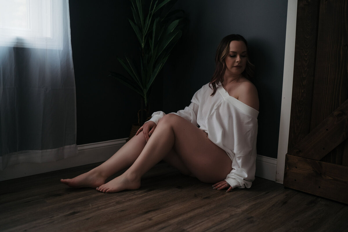A woman in an oversized white long-sleeve shirt sits and leans against on a blue wall by a plant as posed in a New England Boudoir session