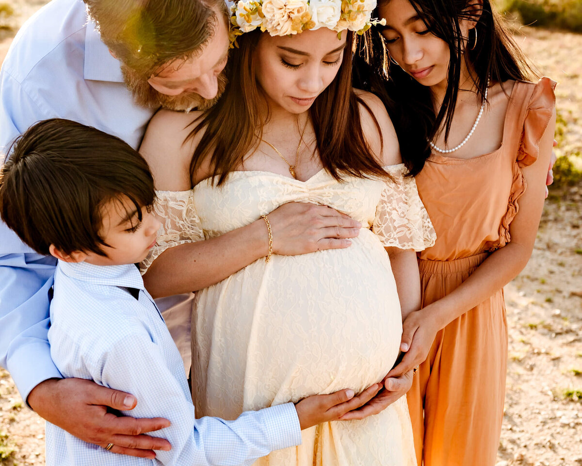 Pregnant mother in cream dress is embraced by her family as they look down at her baby bump